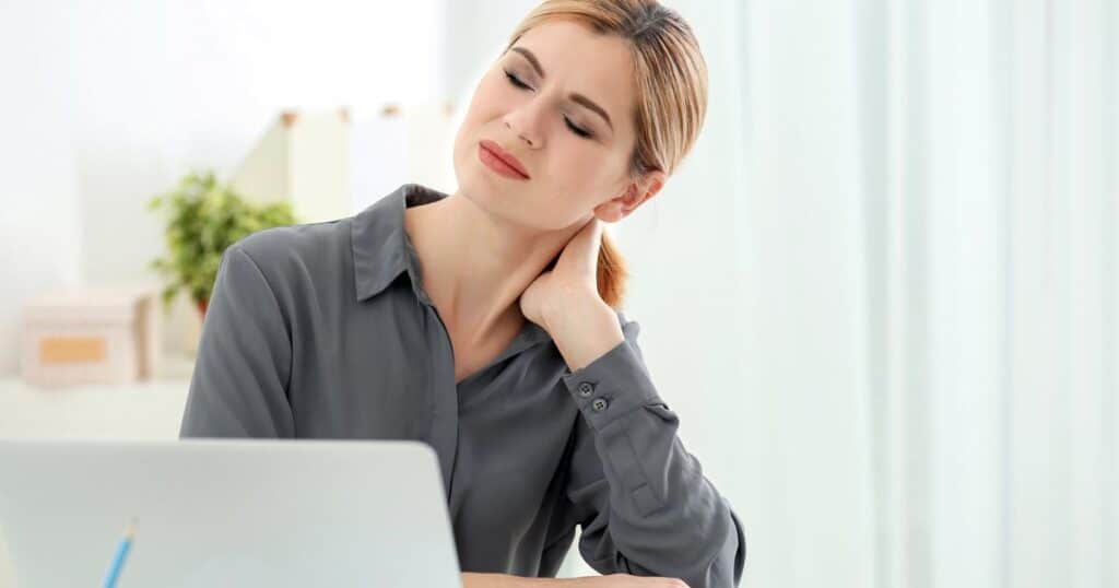 A woman is sitting in front of her laptop touching her neck.