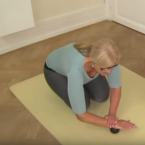 A woman is kneeling on a mat and presses one wrist onto a massage ball.