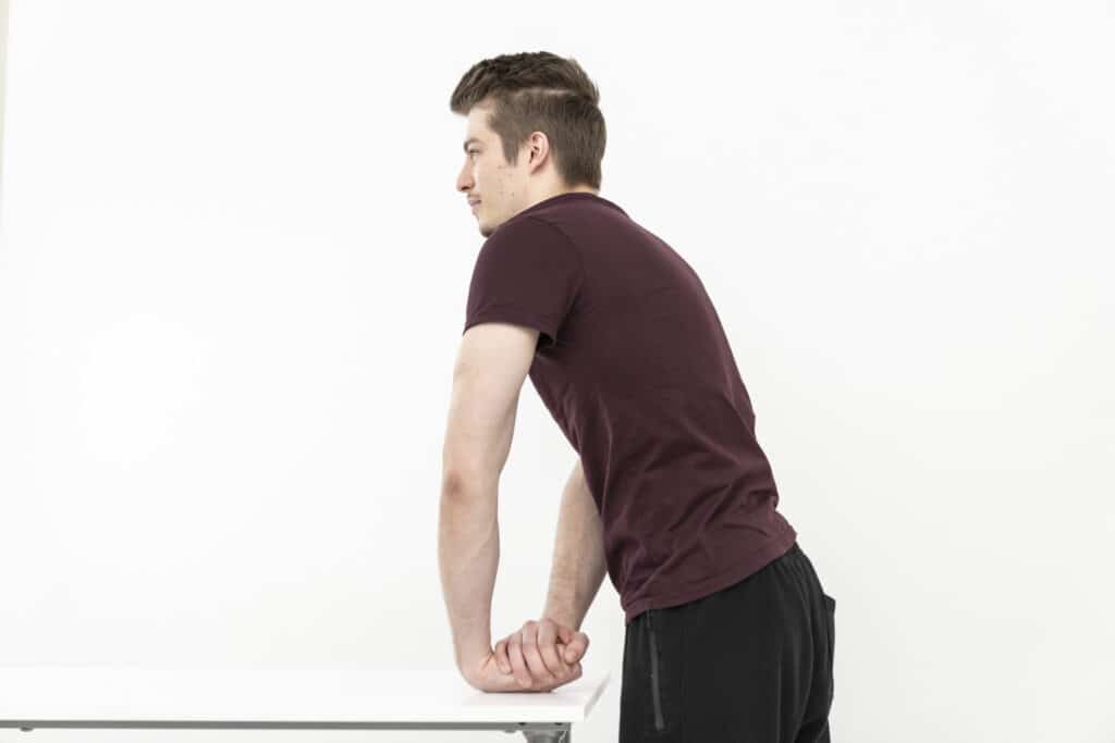 A man stretches his wrist by leaning the outer side of his wrist on the table and straightening his arms.