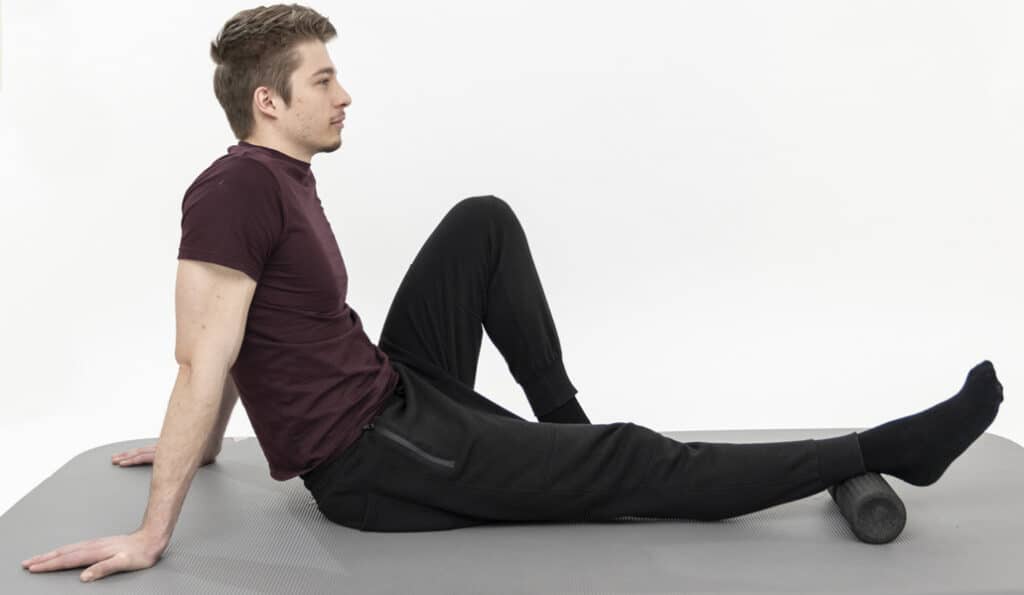 A man is sitting on a mat, stretching out one leg, with the foot propped up on a foam roller. The other leg is angled towards his torso.