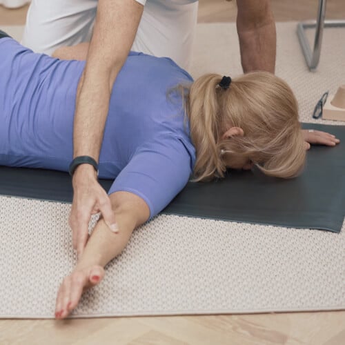A woman lies on the floor with her face down and with her right arm stretched to the side. A man sits on his knees next to her and helps her to stretch the arm.