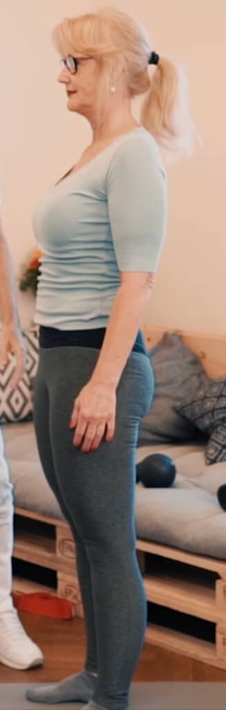 A woman stands up straight holding her arms along her body. There is a sofa in the background.