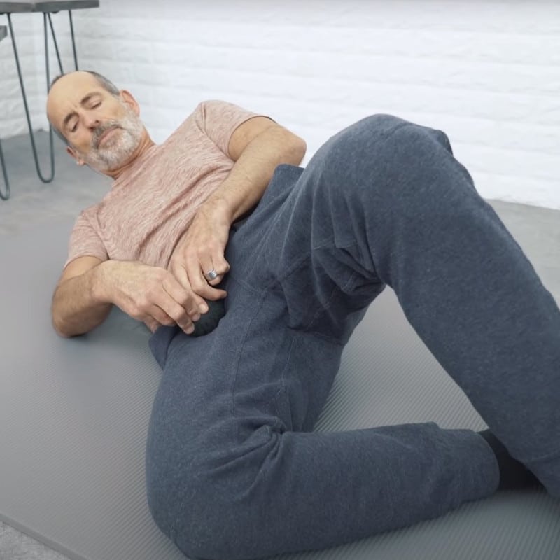 Roland Liebscher-Bracht lies on the ground and presses a massage ball into his right hip. Both legs are bent, the right knee touches the ground, the left knee faces upward.