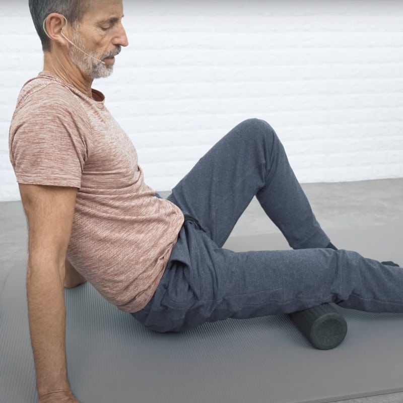 Roland Liebscher-Bracht rolls over a midi foam roller that is placed under his right thigh. The left leg is bent, and both arms are straightened.