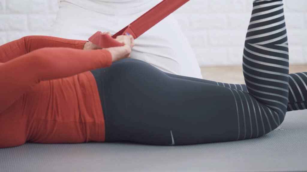 A woman lies face down on the floor and bends her left leg at the knee. She pulls her heel towards the buttlocks using red stretching strap. A man sits on his knees behind her.