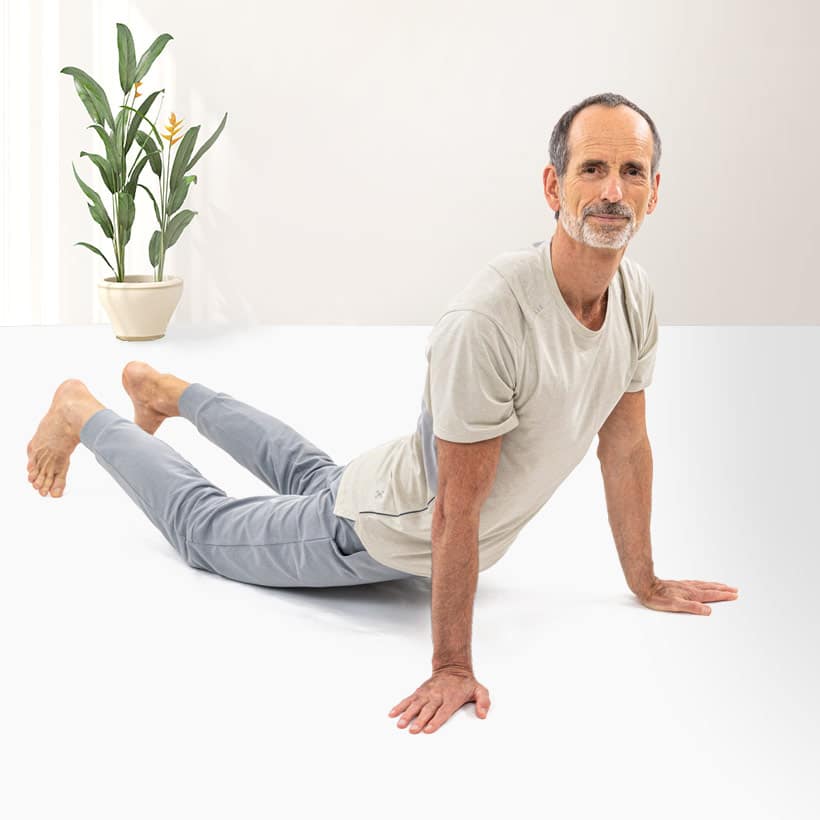 Roland Liebscher-Bracht is doing a stretching exercise on the ground. The lower body touches the ground, arms and upper body are stretched upwards. He looks straight into the camera.