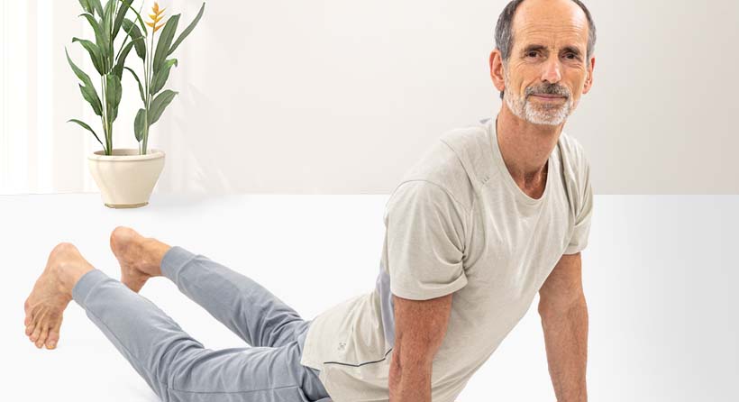 Roland Liebscher-Bracht is doing a stretching exercise on the ground. The lower body touches the ground, arms and upper body are stretched upwards. He looks straight into the camera.