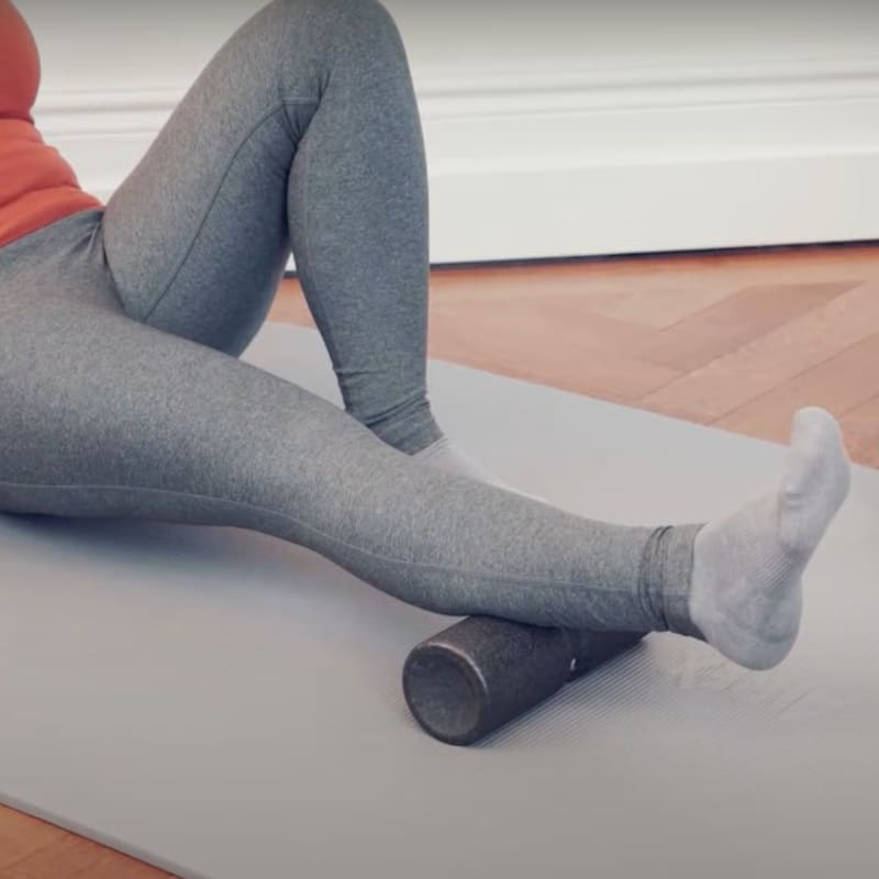 A woman is sitting on the floor with the Midi Foam Roller under her calf.