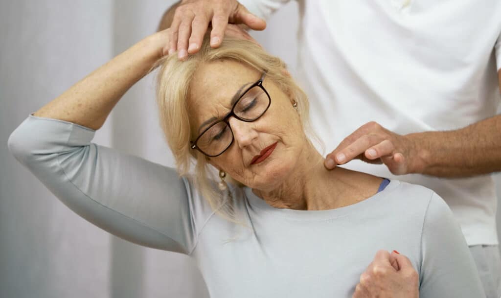 Ina stretches her head to the right side, with her right hand touching her head. The left shoulder pulls downward; her left hand is clenched into a fist. Roland Liebscher-Bracht stands behind her; only his hands and trunk are visible.