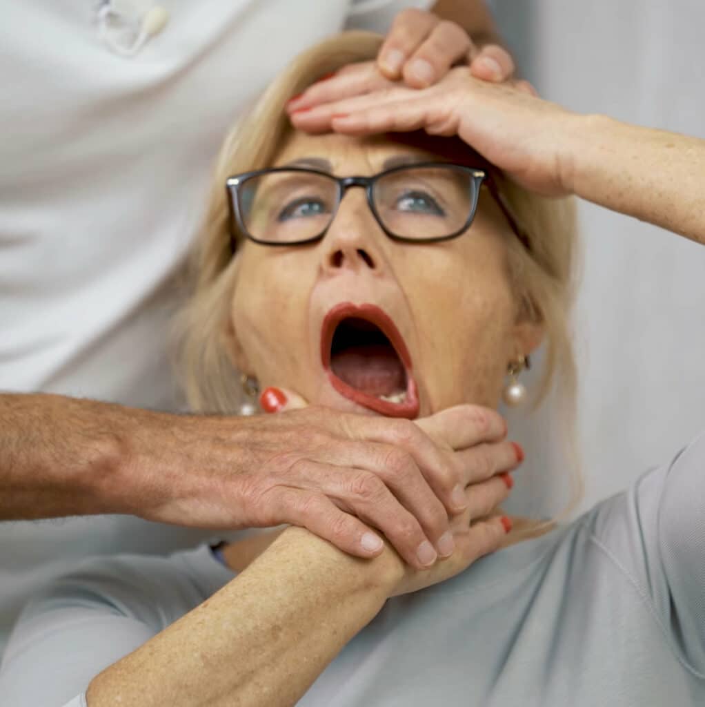 Ina pulls her jaw with the right hand to the left. Her mouth is wide open; her left hand touches the forehead. Two additional hands touch her head and hand.