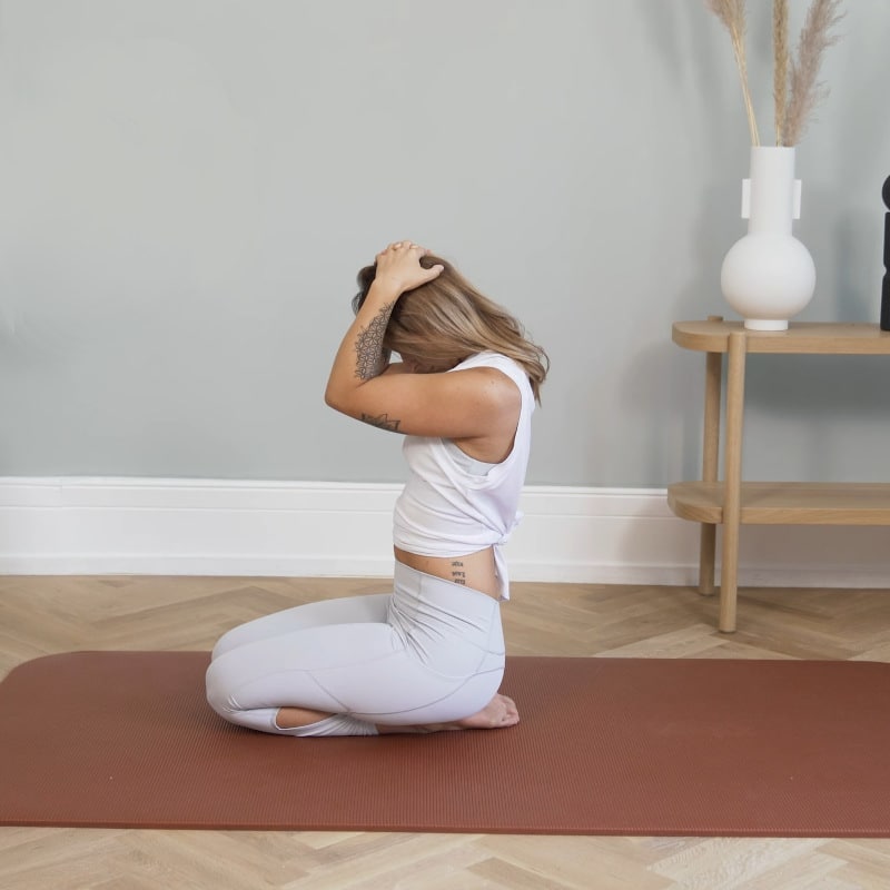 A girl is kneeling on the ground, she's holding her head with both hands, pulling it downwards.
