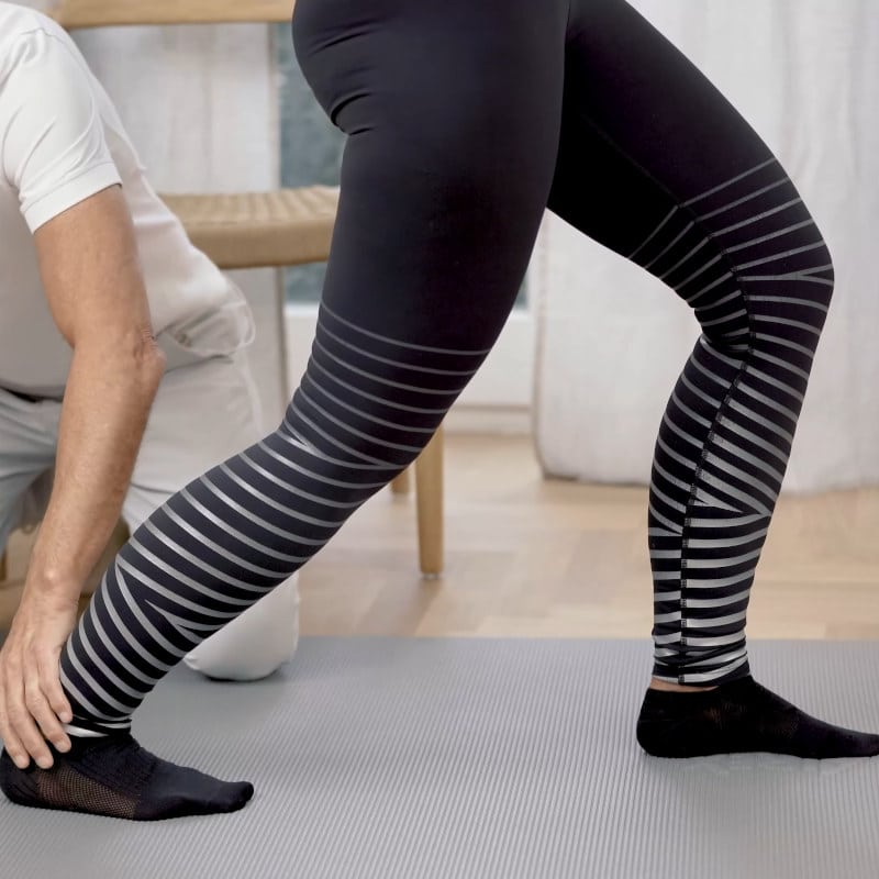 A woman is lunging forward with her left leg and bending slightly at the knees while Roland Liebscher Bracht adjusts her right ankle.