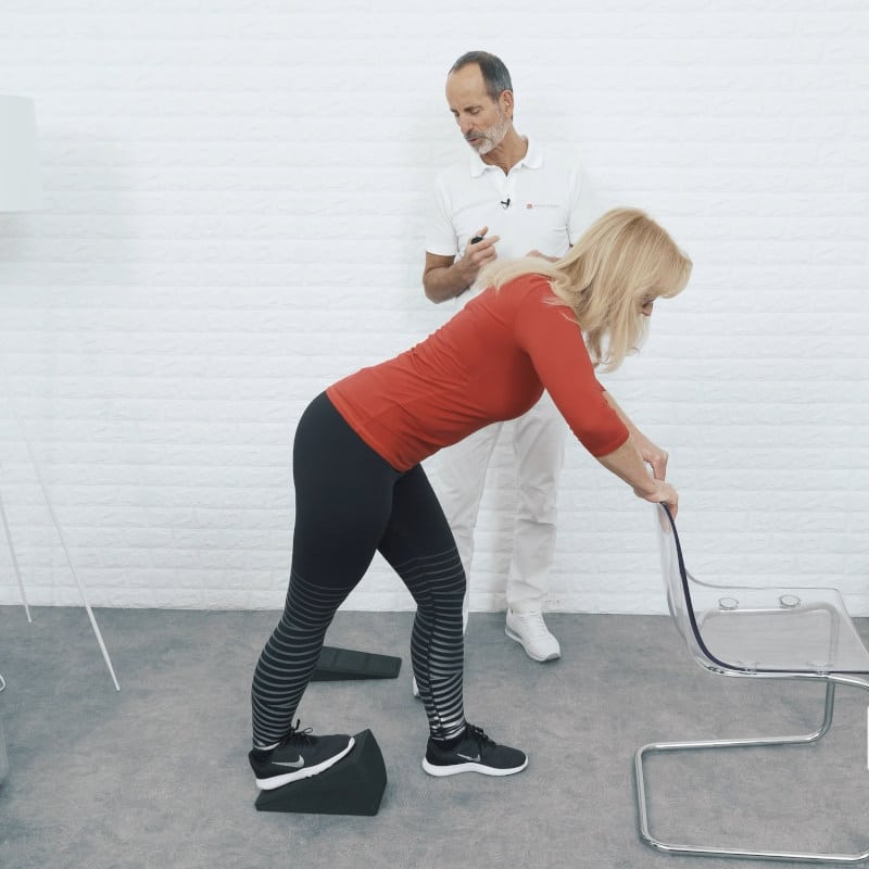 A woman is demonstrating a stretch for Osgood Schlatter pain. She is standing with one foot on the Knee Hero, grabbing the back of a chair while Roland Liebscher-Bracht looks on.
