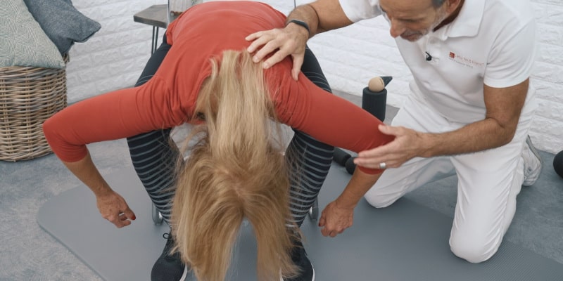 A woman is sitting bent forward between her legs while Roland Liebscher-Bracht looks on.