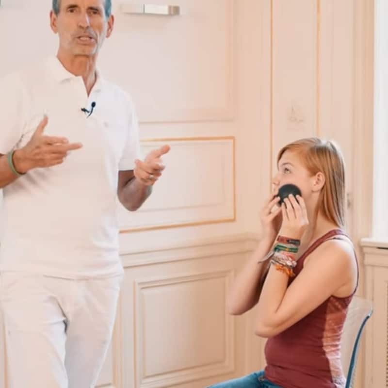 A woman is massaging her left cheek with the Mini Massage ball as Roland Liebscher-Bracht looks on.