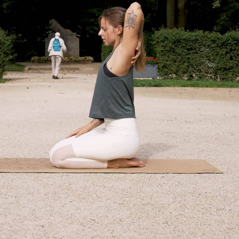 A woman is sitting back on her heels reaching her left arm back touching her shoulder.