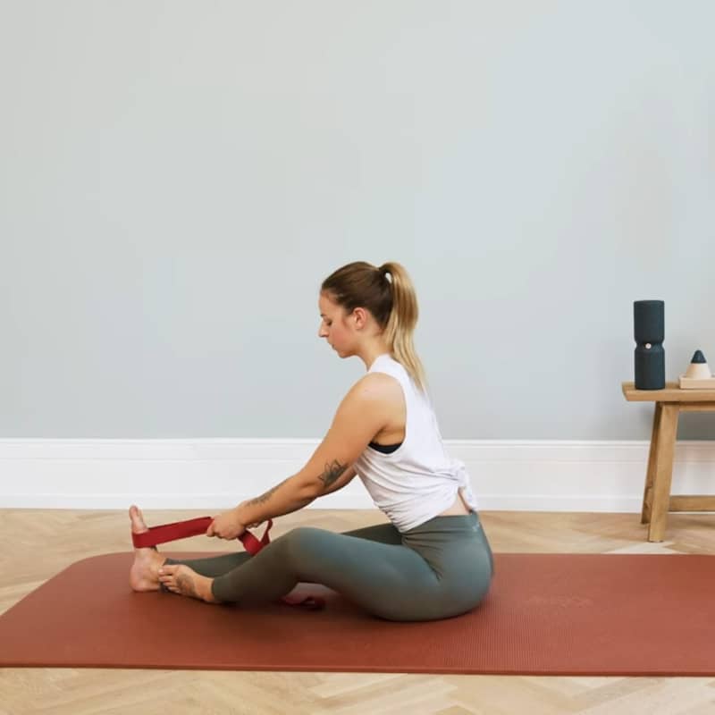 A woman is sitting on the floor with the Liebscher & Bracht Stretching Strap around the ball of her right foot.